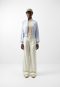 Chaqueta corta de cremallera azul claro con puños acanalados, top acanalado beige, pantalones anchos blancos y gorra beige. Presenta una marca en la chaqueta.
