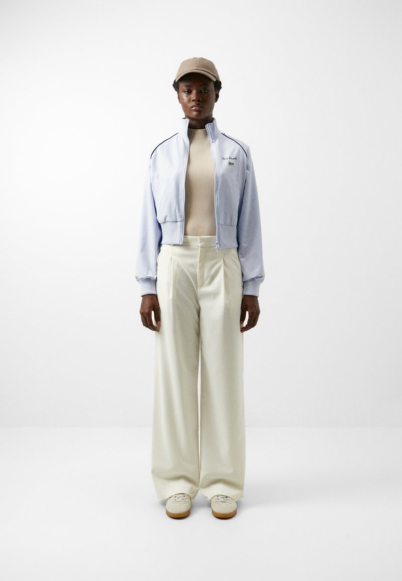 Hellblaues, gekürztes Zip-Jackett mit gerippten Bündchen, beigem, geripptem Oberteil, weißen, weiten Hosen und beigem Hut. Mit Branding auf dem Jackett.