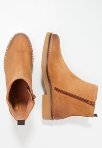 Stivaletti alla caviglia in suede color nocciola con tacco spesso, chiusura con zip laterale e cuciture decorative lungo il bordo. Texture liscia e design snello.