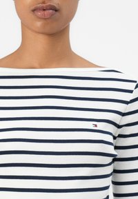 Kvinne som har på seg en hvit båt-hals t-skjorte med svarte horisontale striper og en liten rektangulær rød, hvit og blå logo på brystet.