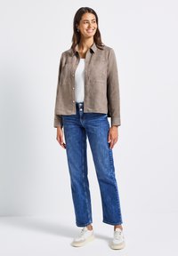 Frau steht und lächelt, trägt eine beige Knopfjacket, ein weißes Oberteil, blaue Jeans und weiße Sneaker vor einem schlichten weißen Hintergrund.