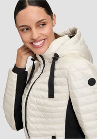 Giacca imbottita di colore off-white con dettagli neri. Presenta un cappuccio, chiusura con zip e cordini regolabili. Design a quadri testurizzati.