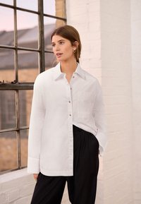 Hvid skjorte med krave, forlommer og let skinnende overflade, parret med sorte, løstsiddende bukser. Simpelt design, lange ærmer.