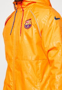 Orange regnjacka med reflexdetaljer, dragkedja fram och huva. Har en Barcelona FC-logga och ett strukturerat mönster på tyget.