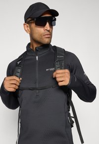 Man in een zwarte pet, met oversized zonnebril, zwarte Columbia zip-up jack, die de banden van een zwarte rugzak aanpast tegen een effen achtergrond.