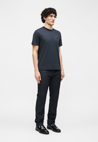 Νεαρός άνδρας με σγουρά μαλλιά, ντυμένος με εφαρμοστό σκούρο μπλε t-shirt, μαύρα τζιν και μαύρα δερμάτινα παπούτσια, στέκεται μπροστά σε ένα απλό φωτεινό φόντο.