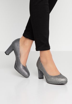 Frau trägt silberne, strukturierte, geschlossene Blockabsatzschuhe kombiniert mit schwarzen eng anliegenden Hosen, steht auf einem glatten, hellen Boden.