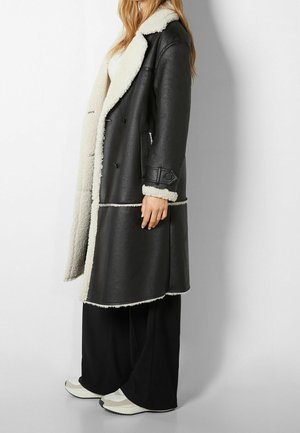 Personne portant un long manteau en peau retournée noire avec une doublure en laine blanche, un pantalon large noir et des baskets blanches, debout de profil contre un mur blanc.