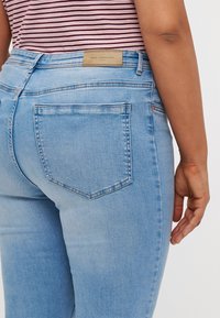 Light blue denim jeans with a mid-rise waist, curved seams, and two back pockets. The brand label is visible at the rear waistband.
