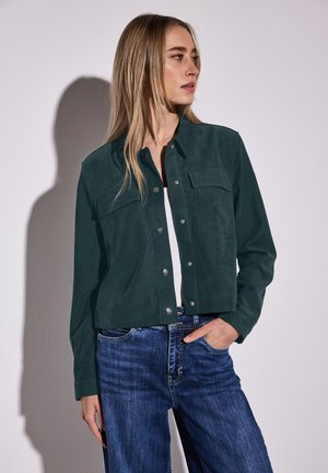Mörkgrön mockajacka med kort modell, knäppning fram, två bröstfickor och texturerad yta, kombinerad med mörka denimjeans.