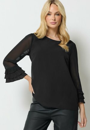 Mujer rubia que lleva una blusa negra transparente de manga larga con puños fruncidos y pantalones de cuero negros, de pie frente a un fondo liso.