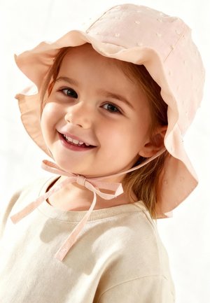 Lächendes junges Mädchen mit einem pinkfarbenen Sonnenhut, der unter dem Kinn gebunden ist, und einem hellbeigen Hemd, das leicht nach links auf weißem Hintergrund blickt.