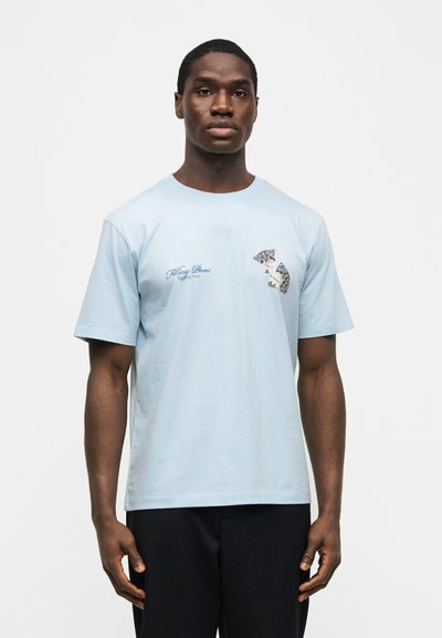 Άνδρας που φοράει ανοιχτό μπλε κοντομάνικο μπλουζάκι με το κείμενο "Filling Pieces" και θρυμματισμένο γραφικό στο στήθος, συνδυασμένο με μαύρο παντελόνι.