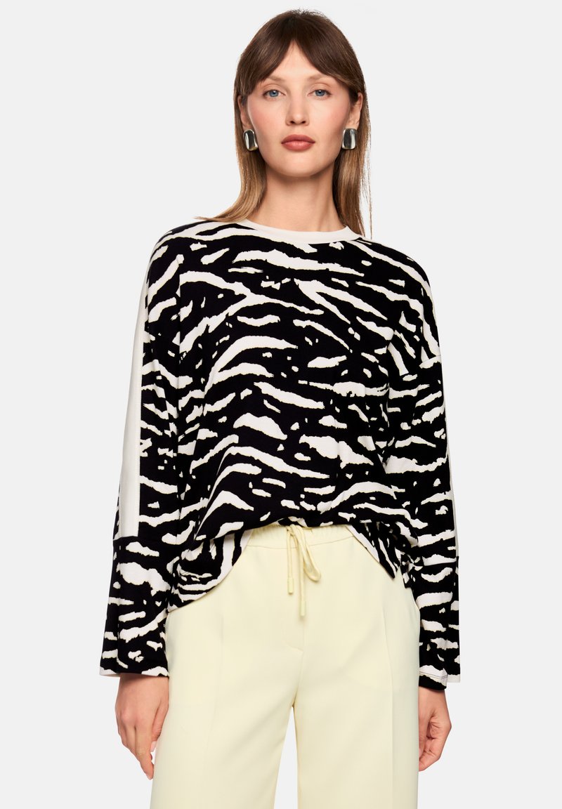 Black and white zebra-striped long-sleeve top with a relaxed fit; paired with light yellow trousers and oversized silver earrings.
