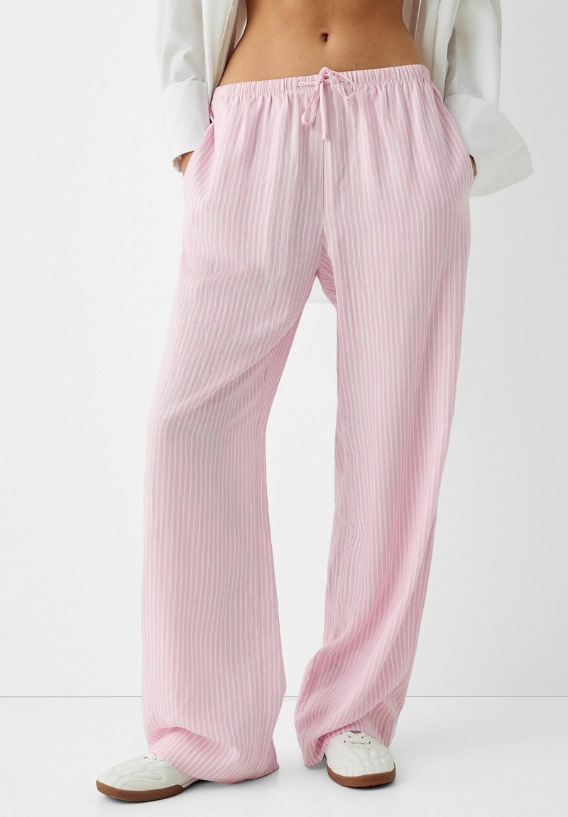 Personne portant un pantalon de pyjama ample à rayures roses et blanches avec cordon, des baskets blanches et une chemise blanche, les mains dans les poches.