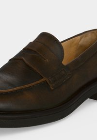 Mocassin en cuir marron avec un bout arrondi, une bride sur le dessus et une semelle noire basse sur un fond clair.