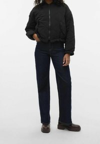 Giubbotto bomber trapuntato nero con chiusura a zip, orlo a coste e tasche laterali, abbinato a jeans a gamba dritta blu scuro e stivali di pelle spessi marroni.
