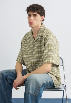 Young man with short curly dark hair wearing green plaid short-sleeve shirt and blue jeans, sitting on a metal chair against plain background.