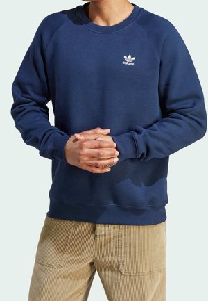 Person trägt dunkelblauen Adidas-Sweatshirt mit Logo auf der Brust und beige Cordhose, Hände vor dem Körper verschränkt.