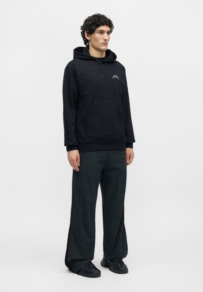 Νέος άνδρας στέκεται, φοράει μαύρη φούτερ με λευκό λογότυπο, φαρδιά μαύρη παντελόνια και μαύρα αθλητικά παπούτσια σε απλό φωτεινό φόντο.