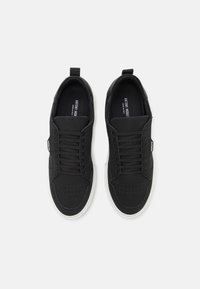 Sneaker noir avec une tige texturée, présentant des perforations près de l'orteil, des lacets plats et une semelle en caoutchouc blanche. Design minimaliste.