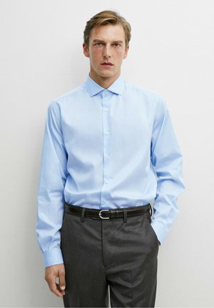 Junger Mann trägt ein hellblaues Hemd, das in graue Hosen mit schwarzem Gürtel gesteckt ist, und steht vor einem schlichten weißen Hintergrund.