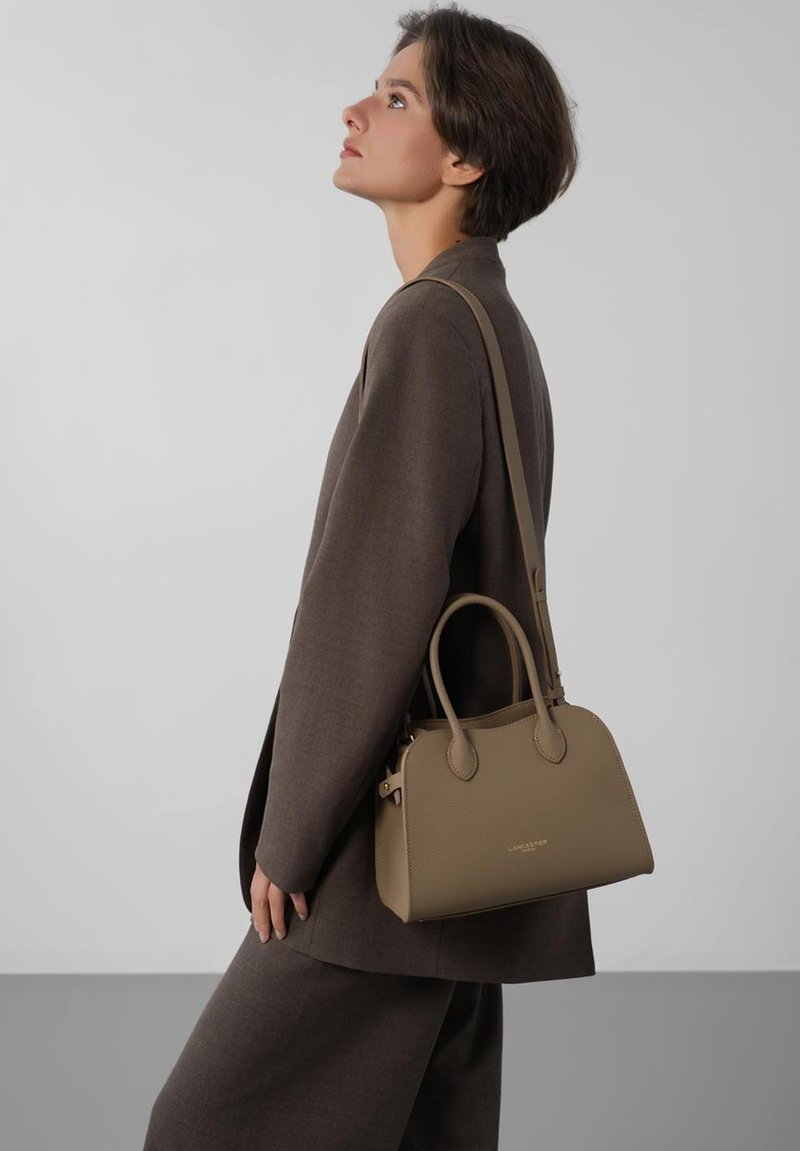 Femme en costume marron portant un sac à main en cuir taupe avec poignées supérieures et bandoulière, regardant vers le haut sur un fond gris uni.