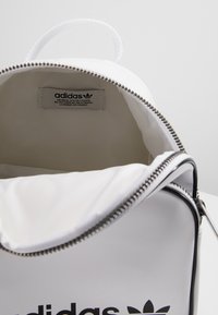 Bílý batoh Adidas s otevřeným zipem, ukazující prázdný interiér a textilní štítek s logem Adidas uvnitř horní klopy.