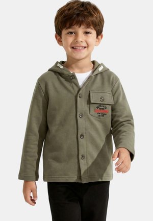 Jeune garçon souriant aux cheveux bruns, portant un sweat à capuche kaki boutonné avec une poche avant et un pantalon noir, debout devant un fond clair.