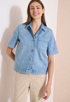 Jeune femme souriante portant une chemise en denim bleu clair à manches courtes et un pantalon beige, debout devant un fond clair bicolore.