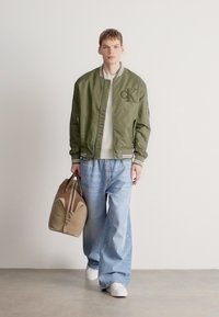 Khaki bomberjakk ribade mansettidega, valge tekstuuriga kampsun, lahtised helesinised teksad ja beež duffelkott. Valged tossud lõpetavad komplekti.