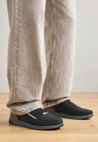 Adidași negri cu un superior texturat, având accente maro și o talpă gri. Purtați cu pantaloni din denim de culoare deschisă, cu o croială lejeră.