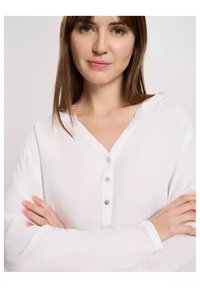 Biała koszula zapinana na guziki z dekoltem w serek i długimi rękawami. Posiada pięć małych guzików na przodzie. Miękka, gładka tekstura materiału.