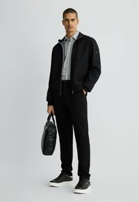 Herrenoutfit mit einer schwarzen Bomberjacke, einem gemusterten Hemd, schwarzen Hosen und Sneakers. Die Accessoires umfassen eine schwarze Lederhandtasche.