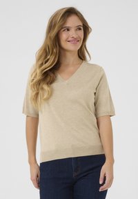 Beige gebreide top met V-hals en korte mouwen; zachte textuur; geribbelde zoom; gecombineerd met donkerblauwe spijkerbroek.