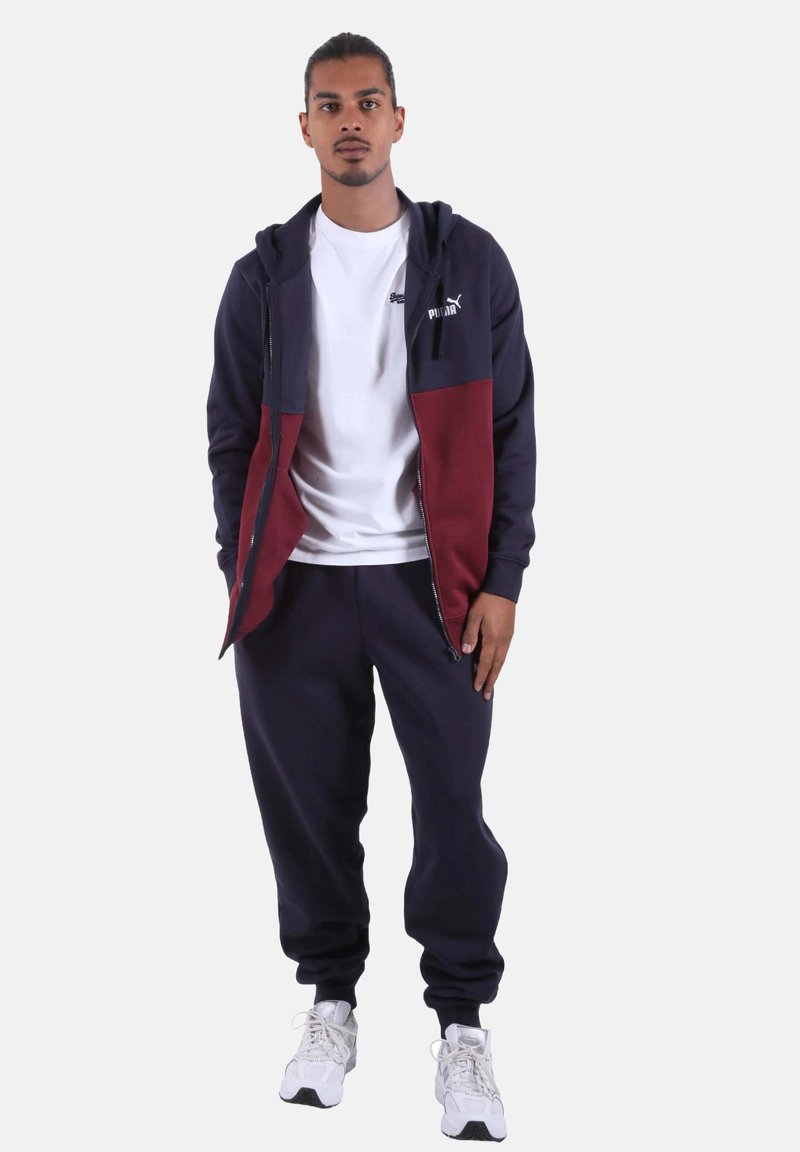 Tuta da uomo in blu navy e bordeaux, realizzata in tessuto morbido. Felpa con cappuccio e zip con logo, abbinata a pantaloni jogger coordinati e sneakers bianche.