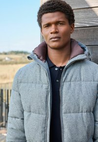 Chaqueta acolchada gris con un collar marrón, hecha de tela texturizada. Lleva puesta sobre una camiseta polo oscura. Fondo natural al aire libre.
