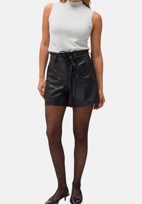 Shorts taille haute en cuir noir avec une ceinture nouée, associés à un haut sans manches gris clair. Le mannequin porte des collants noirs transparents et des chaussures en vernis.