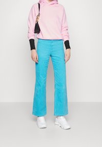 En person klädd i en rosa huvtröja och aqua-blå vida byxor, tillsammans med vita sneakers, står framför en neutral bakgrund.