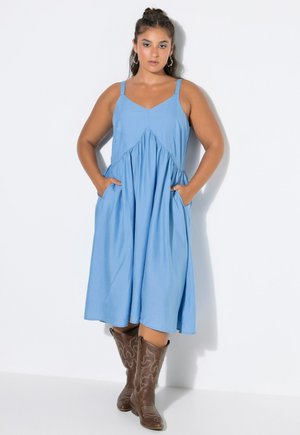 Vrouw met lang donker haar in een blauwe mouwloze midi-jurk met zakken en bruine geborduurde cowboylaarzen staat tegen een witte achtergrond.