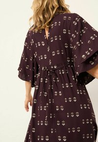 Lila Kleid mit lockerer Passform, kurzen Ärmeln und Taillierungsband. Es hat ein erhabenes gepunktetes Muster in metallischem Gold und Weiß. V-förmiger Ausschnitt am Rücken.