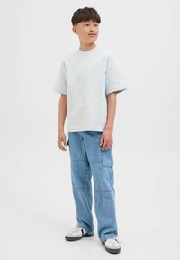 Camicia grigia chiara a maniche corte abbinata a pantaloni cargo blu chiaro con dettagli in patchwork, indossata con sneaker nere e bianche.