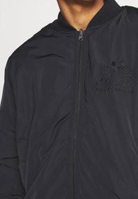 Marinblå bomberjacka tillverkad av slätt tyg, med dragkedja fram, ribbad krage och broderad design med figurer och texten "Håll kontakten."