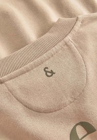 Tessuto di felpa rosa chiaro con collo a costine, caratterizzato da un logo verde "&" e una texture liscia. Primo piano dei dettagli di cucitura e design.