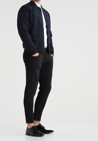 Námořnická bombardérka s kostkovaným vzorem, bílá košile, černé skinny džíny s potrhanými detaily a černé kožené boty.