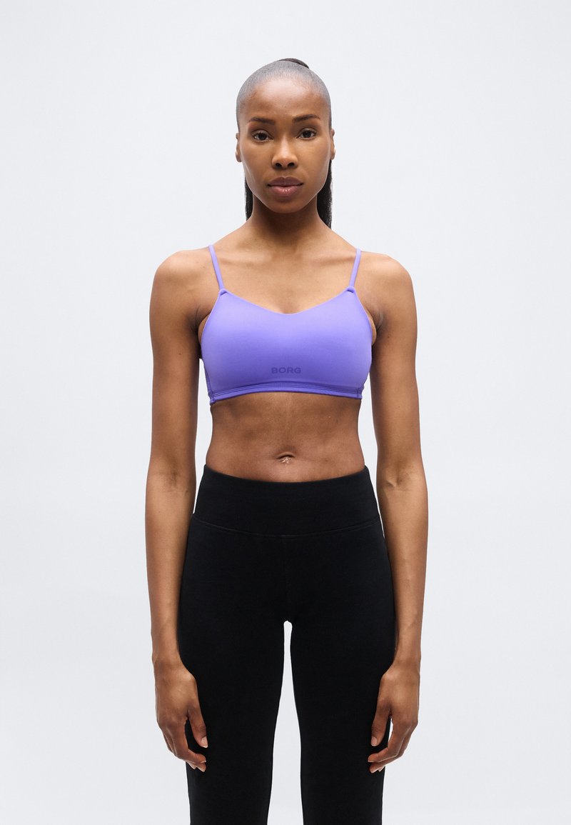 Αθλητική γυναίκα στέκεται μπροστά, φοράει μοβ αθλητικό σουτιέν και μαύρο κολάν με ψηλή μέση, μπροστά σε απλό ανοιχτό φόντο.