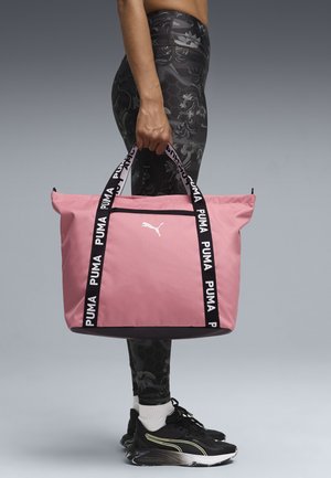 Persona che indossa leggings neri a motivo e sneakers Puma nere, che tiene una grande borsa tote rosa Puma per i manici.