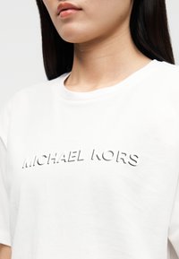 T-shirt branca de algodão com gola redonda, mangas curtas e um logótipo em relevo "MICHAEL KORS" em cor contrastante ao longo do peito.