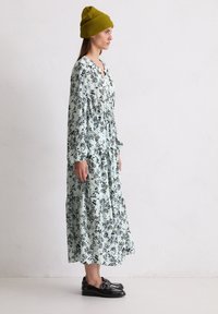 Hellblaues Blumenkleid mit schwarzen Mustern, gestuftem Design, langen Ärmeln und einem knöpfbaren Ausschnitt, kombiniert mit einer gerippten olivgrünen Mütze.
