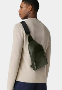 Grüne Leder-Umhängetasche mit einer vorderen Reißverschlusstasche und geprägtlichem Logo, getragen über einem beigen Pullover. Verstellbarer schwarzer Riemen für Komfort.